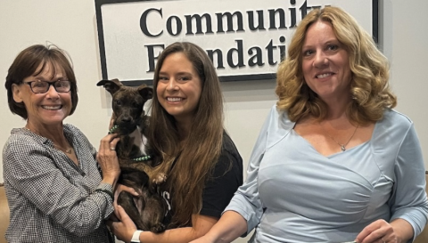 Women smiling with a small dog, standing in front of a foundation sign.